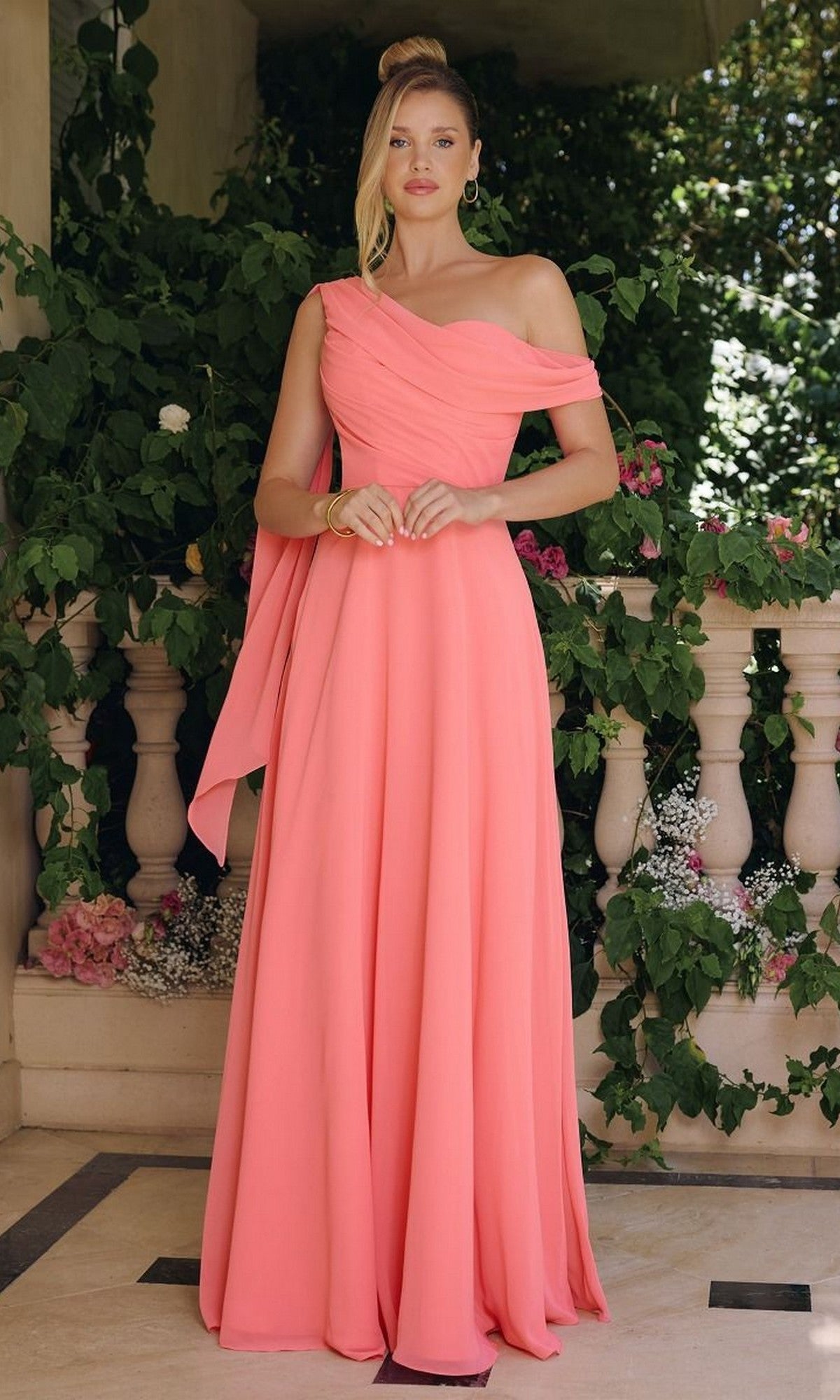 A woman stands before greenery and a stone balustrade wearing the Asymmetric Long A-Line Prom Dress: Amoris T716, a floor-length, off-the-shoulder coral pink gown.